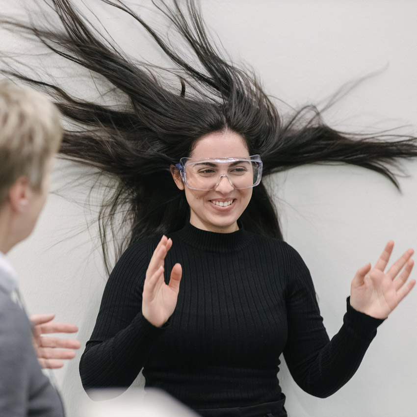 Eine Frau steht im Windkanal an der Wand. Ihr Haar fliegt in alle Richtungen.